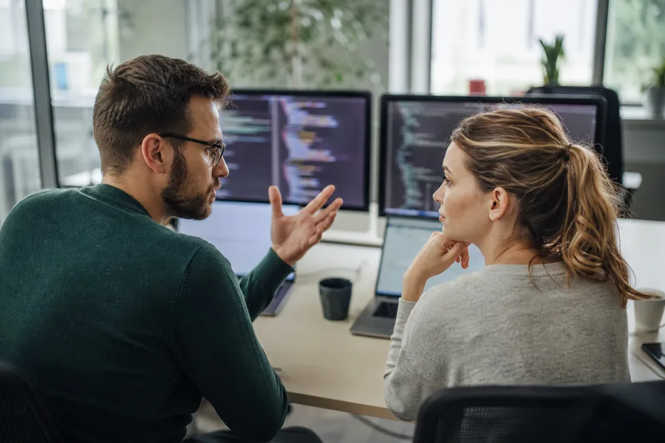 Dwóch programistów w biurze software house omawia kod na dwóch monitorach